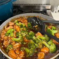 Customer-uploaded image. Click to see more details. The image represents: A stir-fry dish cooking in a yellow-rimmed pan on a stovetop, featuring broccoli florets, chicken pieces, and cashews in a brown sauce, with a black slotted spoon lifting some of the food. A blue pot is visible in the background.