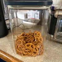 Customer-uploaded image. Click to see more details. The image represents: A clear plastic storage container with a dark blue lid filled with twisted pretzel snacks, sitting on a kitchen counter next to other appliances and items.