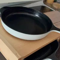 Customer-uploaded image. Click to see more details. The image represents: A white frying pan with black non-stick interior sits on a wooden cutting board stand in a kitchen, with another pan visible below and kitchen appliances in the background