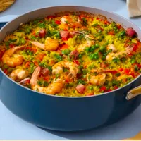 Customer-uploaded image. Click to see more details. The image represents: A colorful paella dish with shrimp, rice, and vegetables served in a large blue pan on a white table with accompaniments