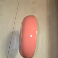 Customer-uploaded image. Click to see more details. The image represents: A coral-pink colored Swiss Army knife or multi-tool with a rounded, closed blade positioned vertically against a beige wallpapered wall with a subtle leaf pattern.