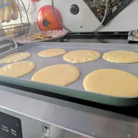 Customer-uploaded image. Click to see more details. The image represents: A baking tray with seven pancakes at various stages of cooking on a stovetop, with kitchen utensils and cookware visible in the background.