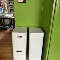Customer-uploaded image. Click to see more details. The image represents: White recycling bins with green lids positioned against a bright green wall in a home interior with wooden floors