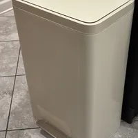 Customer-uploaded image. Click to see more details. The image represents: A white rectangular trash can with a flat lid and foot pedal, positioned on a tiled floor next to a wall and dark furniture.
