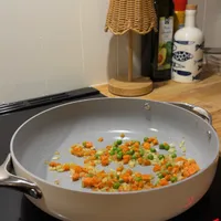 Customer-uploaded image. Click to see more details. The image represents: A white ceramic pan on a stovetop containing diced carrots, peas, and onions being sautéed. Kitchen counter in background with a wicker lamp, wine bottle, and decorative containers.