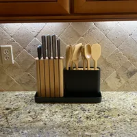 Customer-uploaded image. Click to see more details. The image represents: A black utensil holder on a granite countertop containing kitchen knives with black handles on the left side and wooden cooking utensils on the right side, with a travertine tile backsplash and wooden cabinets visible above.
