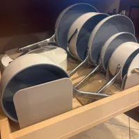 Customer-uploaded image. Click to see more details. The image represents: A set of Caraway brand cookware with cream and dark gray non-stick pans and lids arranged in a wooden drawer organizer.