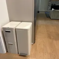 Customer-uploaded image. Click to see more details. The image represents: Two white stackable trash bins with step pedals placed on wooden flooring near a doorway leading to a living room