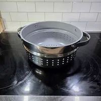 Customer-uploaded image. Click to see more details. The image represents: A stainless steel colander with perforated holes and handles sitting on a black stovetop surface in a kitchen with white subway tile backsplash