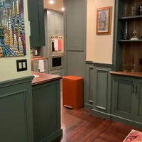 Customer-uploaded image. Click to see more details. The image represents: A modern kitchen interior with sage green cabinetry, stainless steel appliances, wooden countertops with orange trim, recessed lighting, and decorative artwork on the walls including a colorful abstract piece and framed prints. An orange waste bin sits on the dark wood flooring.