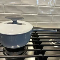 Customer-uploaded image. Click to see more details. The image represents: Blue ceramic saucepan with white interior and lid sitting on black stove grate against white subway tile backsplash