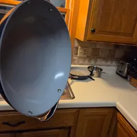 Customer-uploaded image. Click to see more details. The image represents: A large gray oval skillet with a wooden handle hangs from an orange rack above a kitchen counter, with wooden cabinets, a stovetop, and various cooking utensils visible in the background.