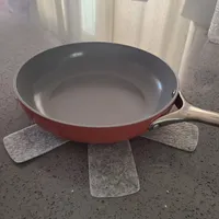 Customer-uploaded image. Click to see more details. The image represents: A red frying pan with a gray non-stick interior and cream-colored handle sits on a dark speckled countertop, surrounded by gray felt pot protectors.