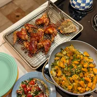 Customer-uploaded image. Click to see more details. The image represents: A home-cooked meal spread on a kitchen counter featuring roasted chicken pieces on a wire rack, a bowl of roasted vegetables with sweet potato and green beans, a blue plate with a fresh tomato and herb salad, and a decorative blue and white checkered bell on the stovetop.
