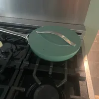 Customer-uploaded image. Click to see more details. The image represents: A green round lid with a metal handle rests on a black pot on a gas stovetop, with a stainless steel range hood visible above.