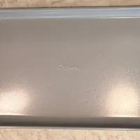 Customer-uploaded image. Click to see more details. The image represents: A rectangular metal baking sheet or cookie sheet with a light gray finish and slightly textured surface, photographed from above on a beige carpet.