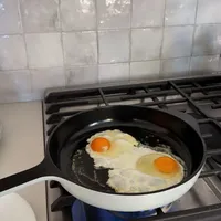 Customer-uploaded image. Click to see more details. The image represents: Two fried eggs cooking in a black and white frying pan on a gas stovetop in a kitchen with white tile backsplash