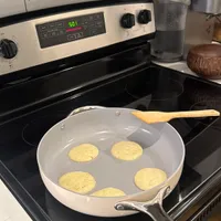 Customer-uploaded image. Click to see more details. The image represents: A white skillet on a black stovetop containing five round, pale yellow pancakes cooking. A wooden spatula rests on the pan's edge. The kitchen has white subway tile backsplash, stainless steel appliances, and a water pitcher visible on the counter.