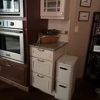 Customer-uploaded image. Click to see more details. The image represents: A kitchen corner showing built-in stainless steel appliances on the left, a white cabinet with granite countertop in the center holding a wicker basket, and a white two-tier trash/recycling bin on the right. Framed artwork and shelving are visible on the tan walls.