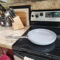 Customer-uploaded image. Click to see more details. The image represents: A kitchen counter scene showing a white ceramic skillet with metal handle resting on a black electric stove cooktop. Behind it are kitchen utensils in a holder, cutting boards, and a knife block. The countertop is granite, and there's a stainless steel range hood and cabinets visible in the background.
