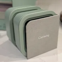 Customer-uploaded image. Click to see more details. The image represents: A mint green Caraway brand compact kitchen appliance with a fabric front panel and rounded rectangular shape, displayed on a light surface.