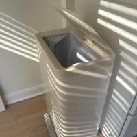 Customer-uploaded image. Click to see more details. The image represents: A white ribbed trash bin with a dark liner bag inside, positioned on a wooden floor next to white blinds in bright sunlight.