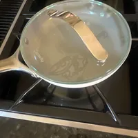 Customer-uploaded image. Click to see more details. The image represents: A glass lid sits on top of a pan filled with clear liquid on a stovetop burner.