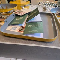 Customer-uploaded image. Click to see more details. The image represents: A yellow tray on a kitchen table displays green and white cookware instruction booklets titled "Clean Cooking Leveled Up," along with product images and a QR code. Yellow cookware and kitchen items are visible in the background.