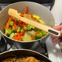 Customer-uploaded image. Click to see more details. The image represents: A person stirring a pot of mixed vegetables including broccoli, red bell peppers, and potatoes with a wooden spoon on a stovetop, with a pot of noodles visible below.