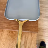 Customer-uploaded image. Click to see more details. The image represents: A square ceramic baking dish with a gold-colored handle sits on a wooden cutting board on a kitchen counter