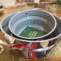 Customer-uploaded image. Click to see more details. The image represents: A stainless steel steamer basket with perforated holes sits on a kitchen counter, with a green book or manual visible inside the basket.