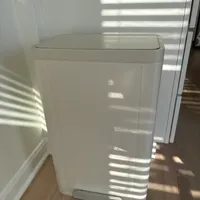 Customer-uploaded image. Click to see more details. The image represents: A white rectangular trash can with a foot pedal at the base, positioned on a wooden floor against a white wall, with sunlight casting shadows across it.