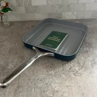 Customer-uploaded image. Click to see more details. The image represents: A square grill pan with ridged bottom and stainless steel handle sitting on a granite kitchen counter, with a green instruction booklet inside the pan