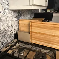 Customer-uploaded image. Click to see more details. The image represents: Kitchen counter with wooden cutting boards stored on a rack above a gas stovetop, with floral patterned wallpaper and white cabinets in the background
