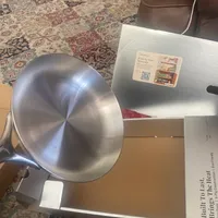 Customer-uploaded image. Click to see more details. The image represents: A stainless steel frying pan sits on top of its original packaging box on a patterned area rug, with a leather chair visible in the background