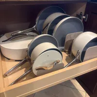 Customer-uploaded image. Click to see more details. The image represents: A kitchen cabinet drawer is open, displaying an organized collection of cookware including cream and gray non-stick pans and pots of various sizes with metal handles, neatly arranged in a wooden drawer organizer.