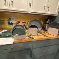 Customer-uploaded image. Click to see more details. The image represents: Kitchen counter displaying organized cookware including green and gray pans with cream-colored lids, arranged on a wooden surface. White cabinets are visible above, and a colorful artist's palette and kitchen items are displayed on the yellow backsplash.