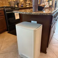 Customer-uploaded image. Click to see more details. The image represents: A modern kitchen with a white rectangular trash can positioned in the center of the floor. The kitchen features dark wood cabinetry, granite countertops, a black stove, and warm lighting from under-cabinet fixtures. A large tumbler and decorative items sit on the island counter in the background.