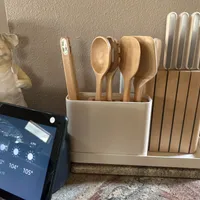 Customer-uploaded image. Click to see more details. The image represents: A kitchen counter display featuring a white ceramic utensil holder with wooden spoons and spatulas, a wooden knife block with metal knives, a decorative white pig figurine, a smart display showing weather information, and a beige textured wall in the background.