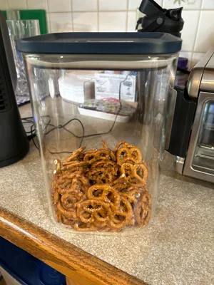 Customer-uploaded image. Click to see more details. The image represents: A clear plastic storage container with a dark blue lid filled with twisted pretzel snacks, sitting on a kitchen counter next to other appliances and items.