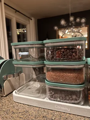 Customer-uploaded image. Click to see more details. The image represents: A set of Caraway brand storage containers with mint green lids arranged on a kitchen counter. The containers hold various baking ingredients including chocolate chips, cocoa powder, and other dry goods. The containers sit on a white base tray in a modern kitchen with dark cabinetry and pendant lighting visible in the background.