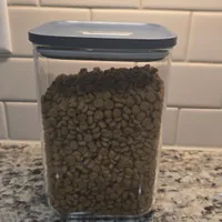 Customer-uploaded image. Click to see more details. The image represents: A clear plastic storage container with a dark gray lid filled with dry pet kibble, sitting on a speckled granite countertop against a white subway tile backsplash.