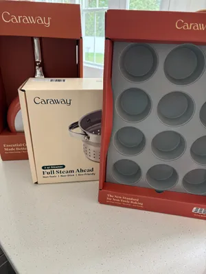 Customer-uploaded image. Click to see more details. The image represents: Caraway brand kitchen products displayed on a counter, including a boxed 3-quart steamer with non-toxic non-stick coating and a muffin tin with multiple circular cups, with red Caraway packaging boxes visible in the background near a window.