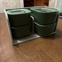 Customer-uploaded image. Click to see more details. The image represents: A set of four green stacked food storage containers with transparent lids, arranged in a white mesh organizer box on a wooden table indoors.