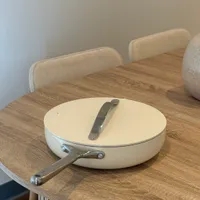 Customer-uploaded image. Click to see more details. The image represents: A cream-colored skillet with lid and metal handle rests on a round wooden table with vertical fluted detailing. Two upholstered chairs with cream-colored backs are visible in the background against a plain white wall.