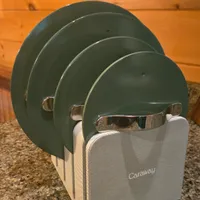 Customer-uploaded image. Click to see more details. The image represents: A set of four nesting green metal bowls with handles stored in a beige Caraway brand fabric container, displayed on a concrete floor against a wooden wall.