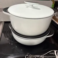 Customer-uploaded image. Click to see more details. The image represents: White ceramic Dutch oven with lid sitting on a black stovetop, with cookbook visible in foreground