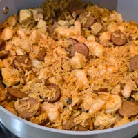 Customer-uploaded image. Click to see more details. The image represents: A white pan filled with paella containing saffron-colored rice, shrimp, sliced sausage, and diced chicken, served on a stovetop.