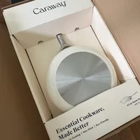Customer-uploaded image. Click to see more details. The image represents: A Caraway brand cookware box is open on a wooden floor, displaying a white saucepan with a stainless steel lid inside. The box shows text reading "Essential Cookware, Made Better" and mentions "Non-Toxic Coating, Non-Stick, Eco-Friendly."