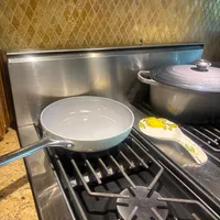 Customer-uploaded image. Click to see more details. The image represents: A kitchen stovetop with a light blue ceramic frying pan on the left burner, a decorative spoon with yellow lemon design, and a dark covered pot on the right burner. Stainless steel backsplash and patterned wallpaper visible in background.